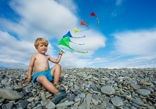 Little boy sit holding many kites in hand on pebble beach over blue sky smiling and looking at camera