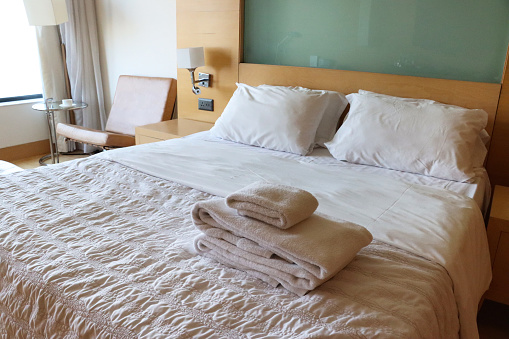 Stock photo showing close-up view of ruck sack in middle of unmade double bed with crumpled, white bedding. In the bedroom beside the bed stands a wooden nightstand and standard lamp.