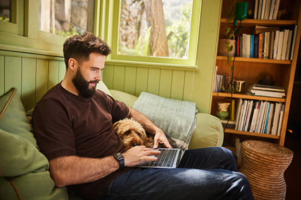 Smiling young man using a laptop on a sofa with his dog