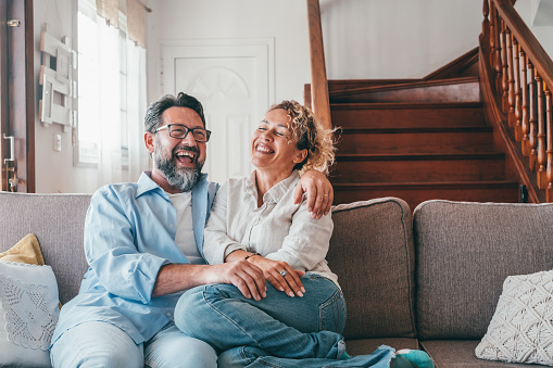 Happy caucasian couple laughing while sitting on sofa in the living room. Husband and wife laughing out loud sitting on couch at home. Man and woman spending leisure time at modern apartment