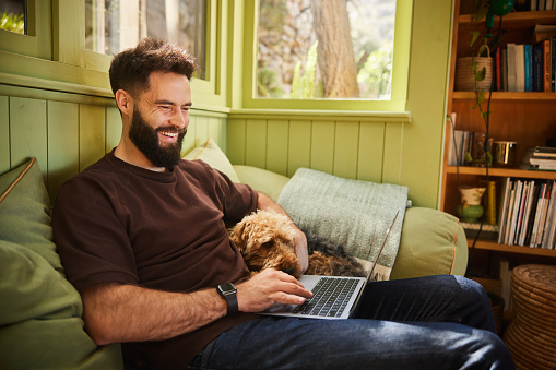 Young man laughing something on a laptop while relaxing with his dog on a living room sofa at home