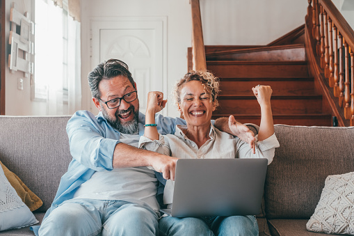 Astonished caucasian couple celebrating online success using laptop sitting on sofa at home. Joyful wife and husband surprised by good unbelievable news, huge shopping sale offer on website