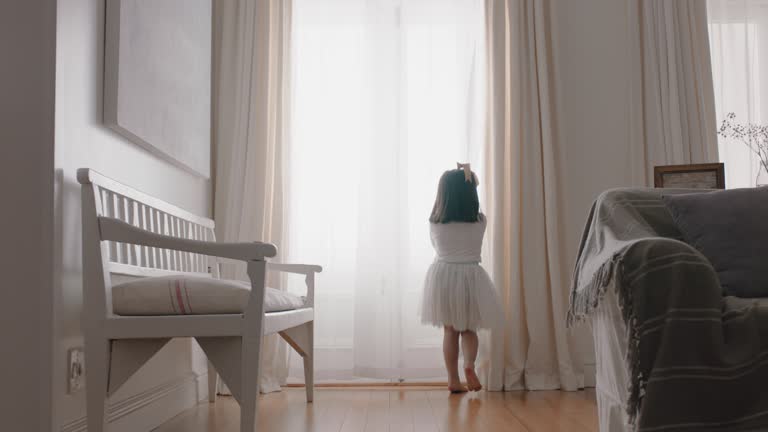 happy little girl running through house wearing tutu opening curtains looking out window at beautiful new day with bright sunlight playful child feeling positive 4k footage