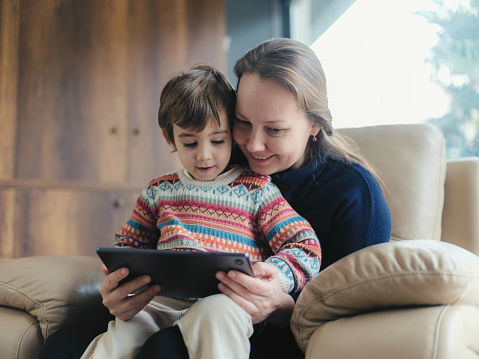 Mother And Little Son Reading Stories For Children On Digital Tablet At Home