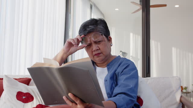 Senior Asian woman with presbyopia, taking off eyeglasses while reading a book at home