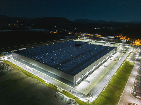 Aerial view of warehouse storage or logistics center from above at night with lights. Industrial distribution warehouse buildings with marked loading docks and trucks waiting to deliver the goods.
