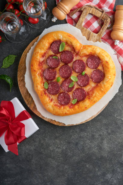 istock Valentines day heart shaped pizza with mozzarella, pepperoni and basil, wine bottle, two wineglass, gift box on black background. Idea for romantic dinner Valentines day. Top view. Mock up. 1464159053