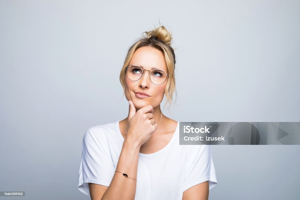 Thoughtful woman with hand on chin looking up Thoughtful blond woman with hand on chin looking up against gray background Contemplation Stock Photo