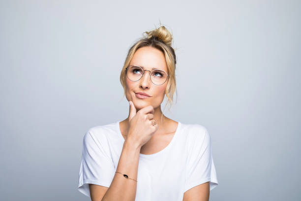 Thoughtful woman with hand on chin looking up Thoughtful blond woman with hand on chin looking up against gray background uncertainty photos stock pictures, royalty-free photos & images