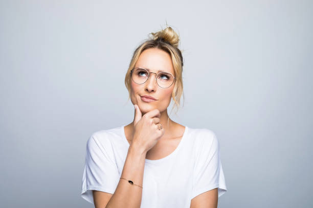 Thoughtful woman with hand on chin looking up Thoughtful blond woman with hand on chin looking up against gray background contemplation stock pictures, royalty-free photos & images