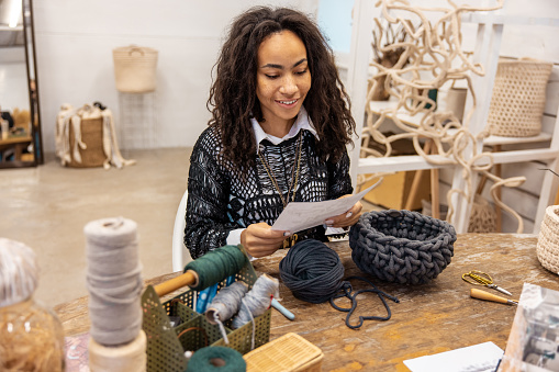 Waist-up portrait of a smiling macrame artist looking at the sheet of paper in her hands