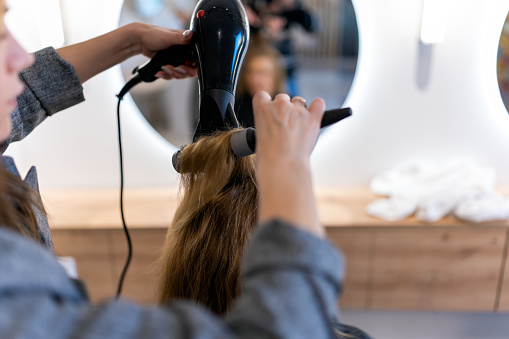 Unrecognizable hairdresser drying hair of anonymous female client with hairdryer and brush in modern hairdressing salon