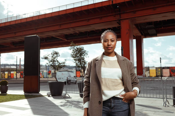 Black woman in town Portrait of a beautiful African-American woman with her hair in braids, wearing a  brown coat and casual clothes posing with the bridge in the background. georgijevic stock pictures, royalty-free photos & images