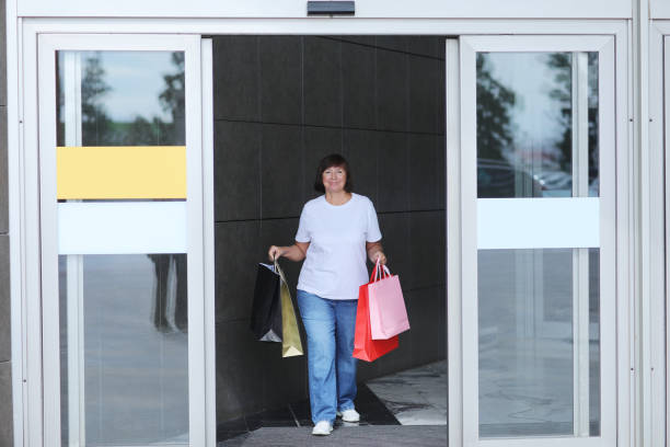 Smiling middle aged woman in casual clothes is walking with colourful shopping bags from shopping mall during Black Friday shopping process, consumerism concept, sale, Season winter summer sales Smiling middle aged woman in casual clothes is walking with colourful shopping bags from shopping mall during Black Friday shopping process, consumerism concept, sale, Season winter summer sales. walking into glass door stock pictures, royalty-free photos & images