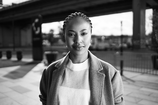 Black and white portrait of a beautiful African-American woman with her hair in braids, wearing a sweater and coat. She is in the town on a cold but sunny day.