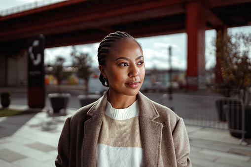 A portrait of a beautiful African-American woman with her hair in braids, wearing a sweater and coat. She is in the town on a cold but sunny day.