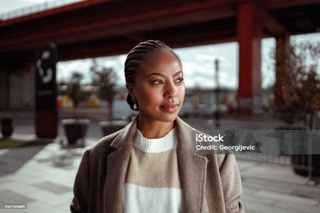 Black woman in town A portrait of a beautiful African-American woman with her hair in braids, wearing a sweater and coat. She is in the town on a cold but sunny day. Portrait Stock Photo