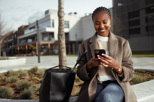 Shot of a beautiful African-American woman using a smartphone in an urban area. She sits on a bench in smart casual clothes on a cold sunny day and smiles.
