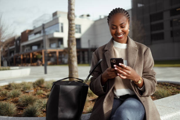 Black woman in town Shot of a beautiful African-American woman using a smartphone in an urban area. She sits on a bench in smart casual clothes on a cold sunny day and smiles. georgijevic stock pictures, royalty-free photos & images