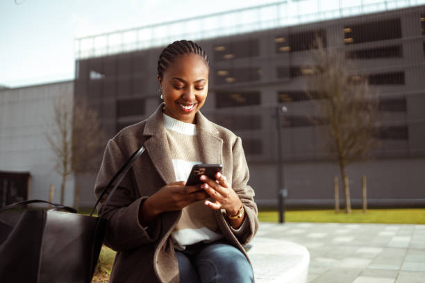 Black woman in town Shot of a beautiful African-American woman using a smartphone in an urban area. She sits on a bench in smart casual clothes on a cold sunny day and smiles. georgijevic stock pictures, royalty-free photos & images