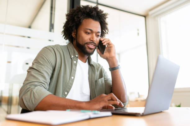 istock Young African-American man using laptop on the workplace and has phone conversation 1464167298