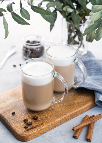 Two cappuccino or latte with milk foam in glasses on a light blue background with coffee beans, cinnamon and eucalyptus branch. The concept of spring breakfast.