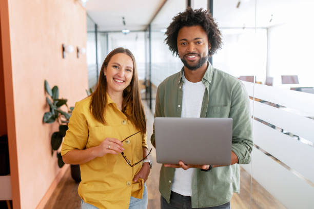 Two happy diverse colleagues African American man and Caucasian woman looking at camera standing in office lobby hall stock photo