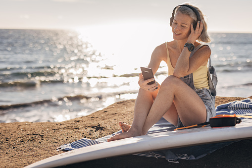 One woman, beautiful female surfer using smart phone and listening music on headphones on the beach by the sea.