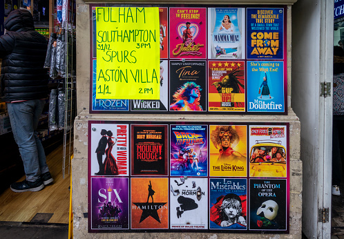 Rows of theatre posters (with a poster for two football matches) outside a bookings agency in London’s West End. The posters include: & Juliet, Mamma Mia!, Come From Away; Frozen, Wicked, Tina, Get Up Stand Up!, Mrs. Doubtfire, Pretty Woman, Moulin Rouge!, Back to the Future, The Lion King, Only Fools and Horses, Six, Hamilton, The Book of Mormon, Les Miserables and The Phantom of the Opera.