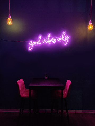 Good vibes only pink neon script over a table and two chairs from the window of a  restaurant