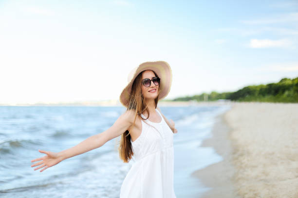 Happy smiling woman in free happiness bliss on ocean beach standing with a hat, sunglasses, and open hands. Portrait of a multicultural female model in white summer dress enjoying nature during travel holidays vacation outdoors. feliz mujer sonriente en felicidad gratuita en la playa del océano de pie con un sombrero, gafas de sol y manos abiertas. retrato de una modelo femenina multicultural en vestido blanco de verano disfrutando de la naturaleza durante las vacaciones de viaje