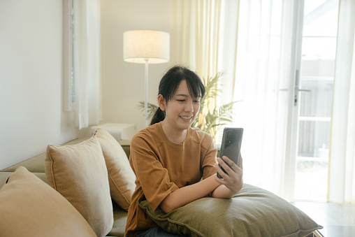 Asian woman enjoying video conversation via smartphone with eldery mother while sitting on sofa during self isolation for coronavirus prevention.
