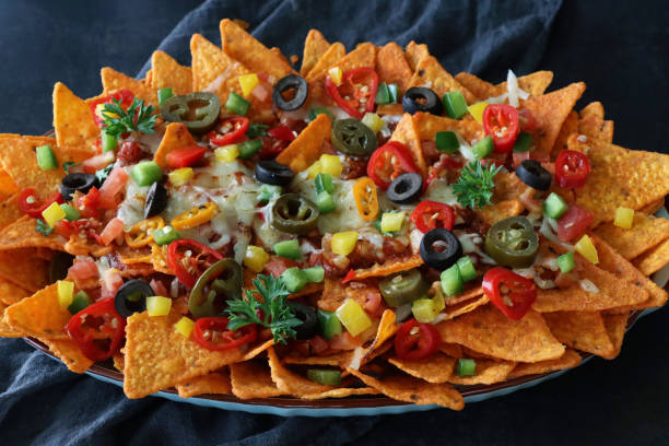 Close-up image of homemade, loaded nachos covered in melted mozzarella cheese, cherry tomatoes, black olives, Jalapeno peppers, grey muslin, black background, elevated view, focus on foreground stock photo