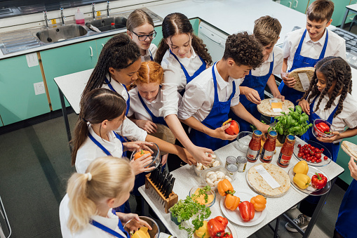 A high angle view of students choosing ingredients as they make pizza in a cooking class in the Northeast of England. They are wearing aprons to keep their school uniforms clean they are using lots of fresh ingredients to cook with. They are working in pairs to make their pizzas.