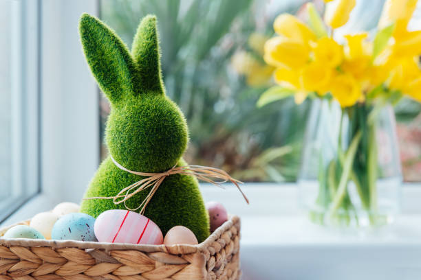 istock Easter bunny rabbit statuette in straw basket with colored eggs on the windowsill with fresh spring tulips and daffodils flowers bouquet on the background. Happy Easter. Selective focus. Copy space. 1464182590