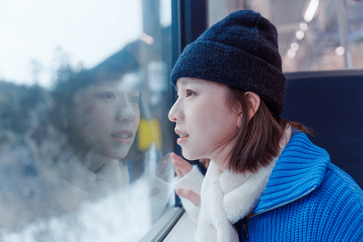 Half-length waist-up side shot of young smiling Asian adult woman, wearing warm winter clothing, looking at the nature landscape snowy mountain views through the window, while sitting in a train.

Young happy Asian female traveler or tourist having fun and relaxing on a solo-trip or travel in Japan during winter holidays, enjoying her day outdoors.