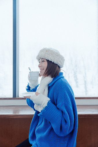 Half-length waist-up side shot of young smiling Asian adult woman, wearing warm winter clothing, holding a cup and drinking hot tea or hot coffee, feeling relaxed while sitting indoors in a cafe in the mountain or park during heavy snow.\n\nYoung happy Asian female traveler or tourist having fun and relaxing on a solo-trip or travel in Japan during winter holidays, enjoying her day outdoors.