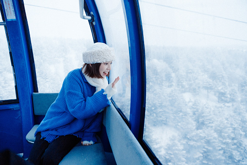 Half-length waist-up side shot of young smiling curious Asian adult woman, wearing warm winter clothing, riding a cable car to the mountain peak while sitting and looking at the nature view or landscape in heavy snow through the window.

Young happy Asian female traveler or tourist having fun and relaxing on a solo-trip or travel in Japan during winter holidays, enjoying her day outdoors.