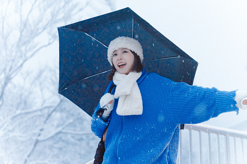 Half-length waist-up shot of young smiling Asian adult woman, wearing warm winter clothing, standing under umbrella and looking at the snowy landscape while exploring the mountain.\n\nYoung happy Asian female traveler or tourist having fun and relaxing on a solo-trip or travel in Japan during winter holidays, enjoying her day outdoors.