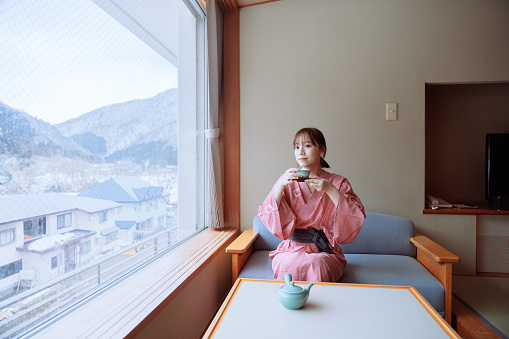 Wide shot of young smiling Asian adult woman, wearing Kimono, sitting and drinking hot tea while looking at the views or landscape of the snowy mountain nature through the big window. She is relaxing inside a traditional Japanese room.\n\nYoung happy Asian female traveler or tourist relaxing during vacation or travel in Japan in Winter holidays, enjoying her day resting indoors.