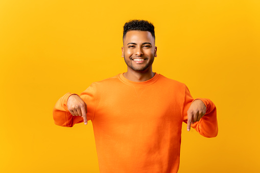 Look, advertise below. Cheerful indian guy pointing down place for commercial idea, looking at camera with toothy smile. Studio shot isolated on yellow background