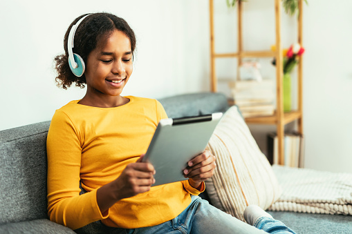 Happy African young girl using digital tablet technology sitting on couch at home. Smiling young girl using apps, make video call, browsing internet on sofa.