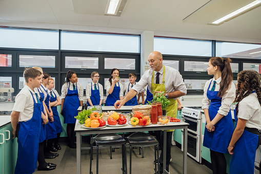 A close up side view of a group of teenage high school students in the North East of England. They are being taught about the nutritional value of the veg they are going to be using on the pizzas that they are about to make. The teacher is advising them how to combine flavours