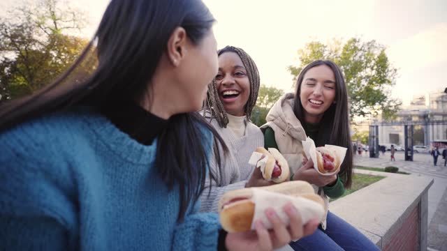 Cheerful Girls Eating Take Away Street Food. Three Happy Girlfriends taking hot dogs in the Park