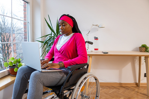 Young woman of Black ethnicity in wheelchair working on laptop, from her home office near window