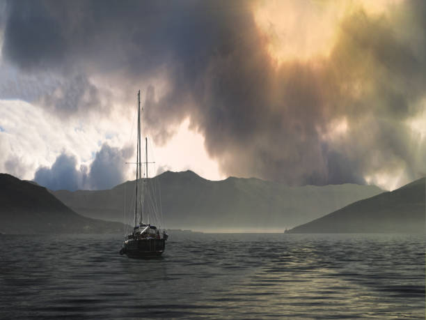 Sea yacht with high mast in water with small waves under dark cloudy sky with visible yellow sun lights and foggy mountains at background back view