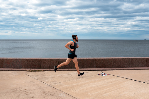 Focused bearded man running outdoors while listening music with headphones near the sea.