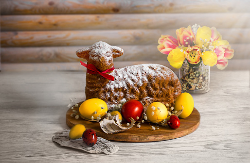 Lamb of God pastry with bright yellow and red eggs, birch bark and a bouquet of tulips in the background. Easter table and treats
