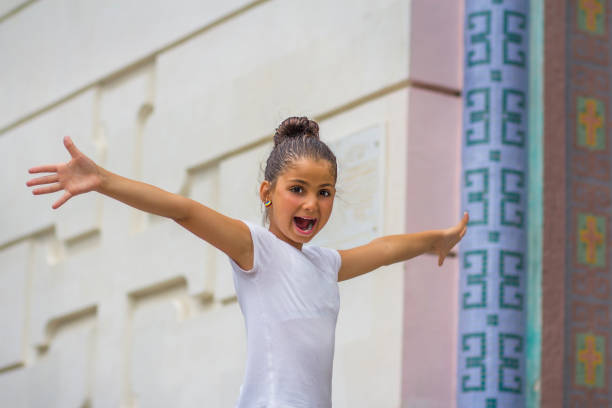 una adolescente de 10 años de apariencia caucásica con una camiseta blanca extiende alegremente sus brazos y grita con admiración y belleza en la entrada de la iglesia contra el fondo de una pared de luz con una cruz. - armenian ethnicity fotografías e imágenes de stock