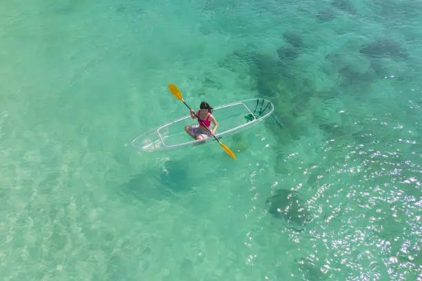 Photo of Aerial top view of Asian woman, a tourist, paddling a boat, canoe, kayak or surfboard with clear blue turquoise seawater, Andaman sea in Phuket island in summer season, Thailand. Water in ocean